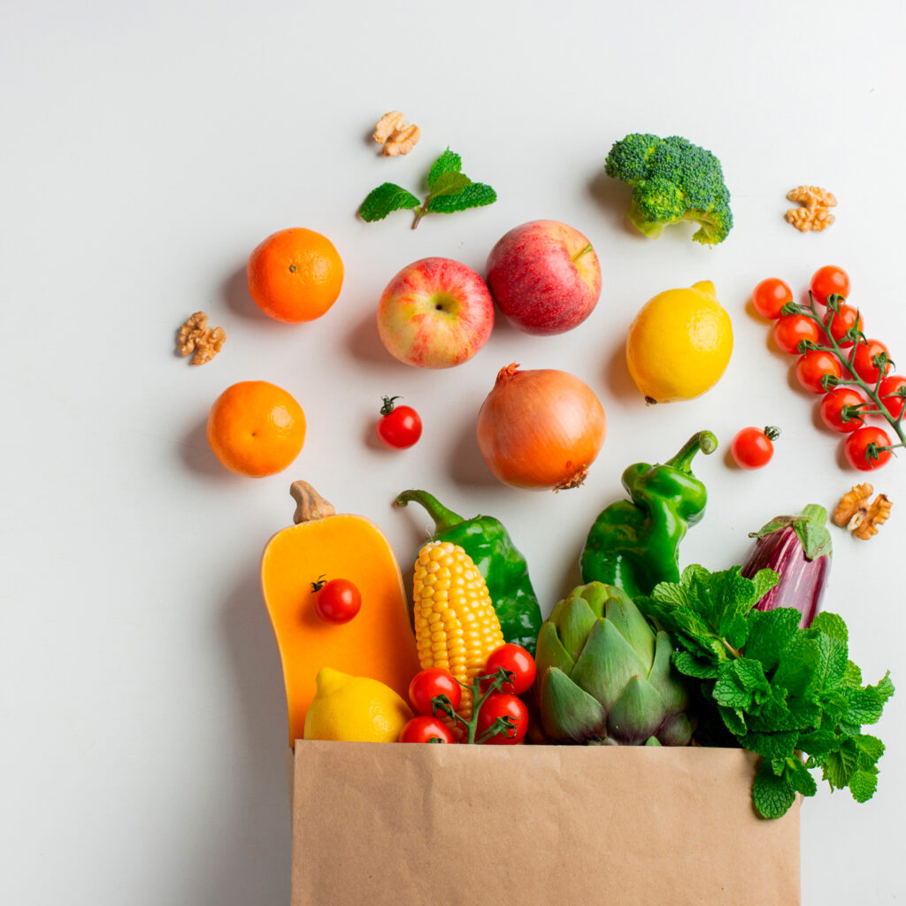 Sacolinha de papel com vegetais e frutas frescas saindo para fora, incluindo milho, pimentão, alcachofra, tomate, maçã e cítricos, sobre fundo branco.