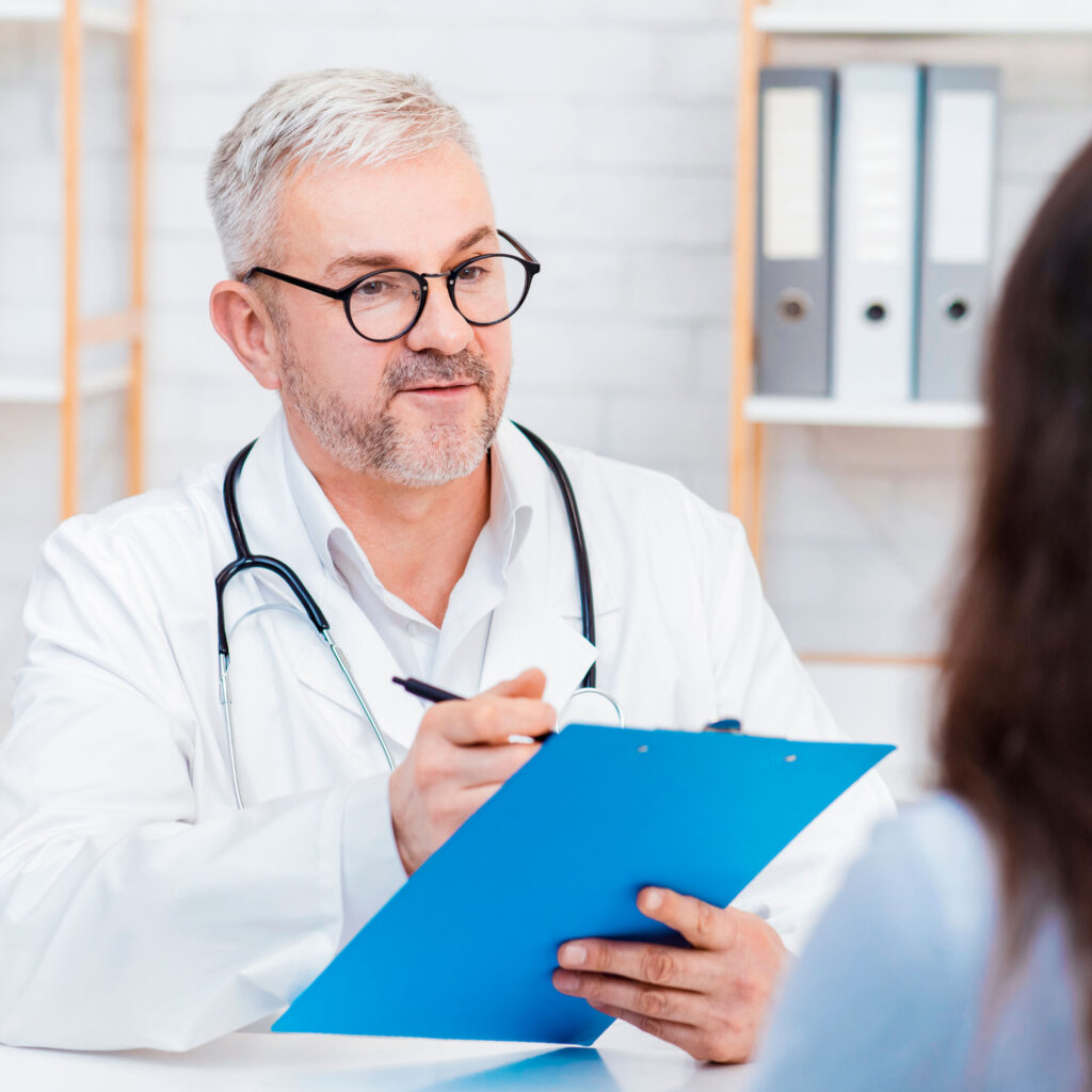 Médico com jaleco e estetoscópio segura uma prancheta azul e conversa com paciente em consultório.