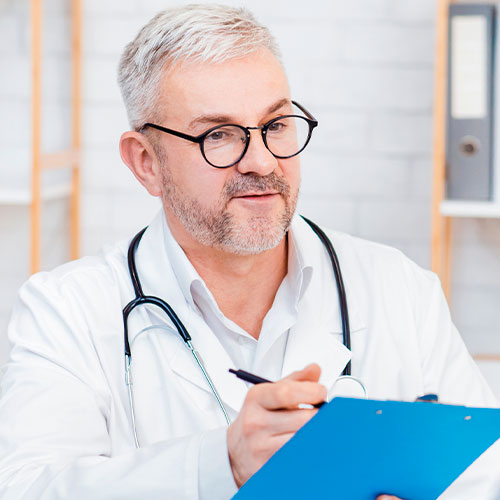 Médico com jaleco e estetoscópio segura uma prancheta azul e conversa com paciente em consultório.