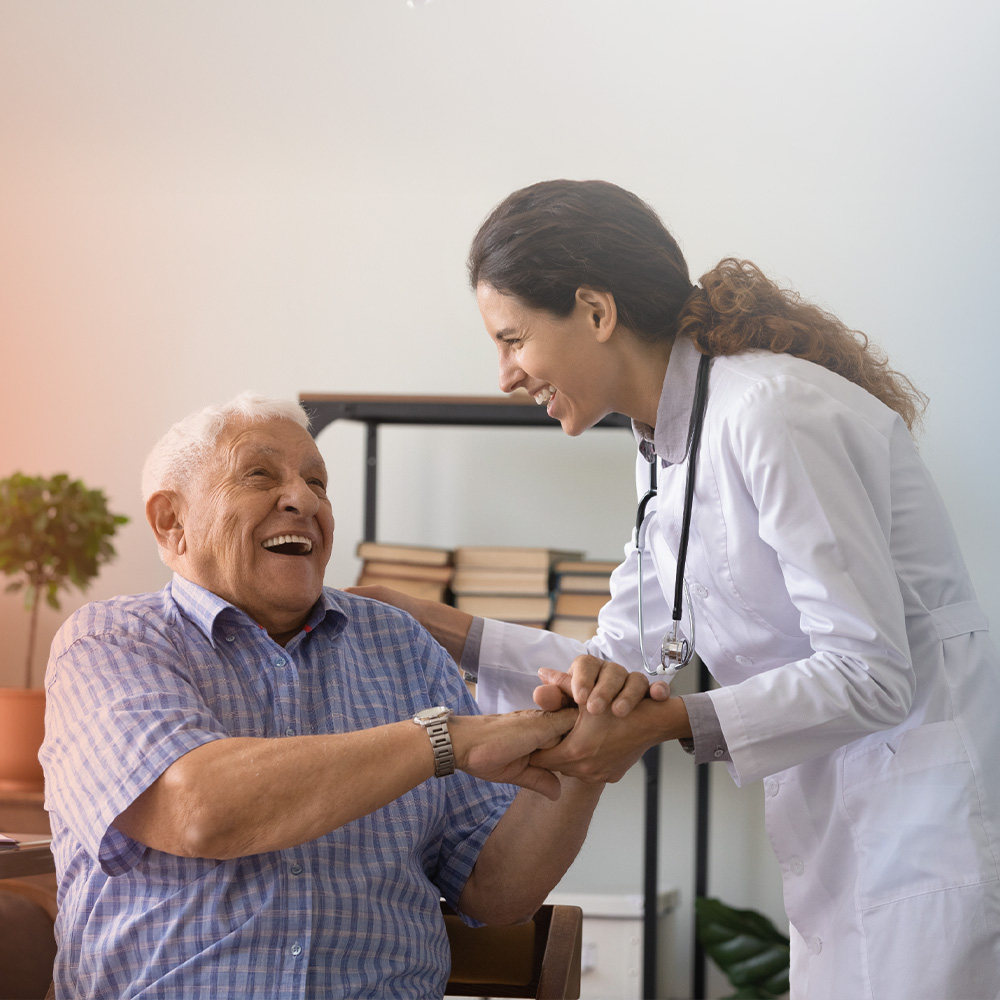 Médica sorridente segura a mão de um idoso também sorridente, em ambiente acolhedor com estante de livros ao fundo.