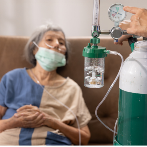 Idosa com máscara de oxigênio sentada em sofá enquanto cilindro de oxigênio é ajustado por um profissional de saúde ao seu lado.