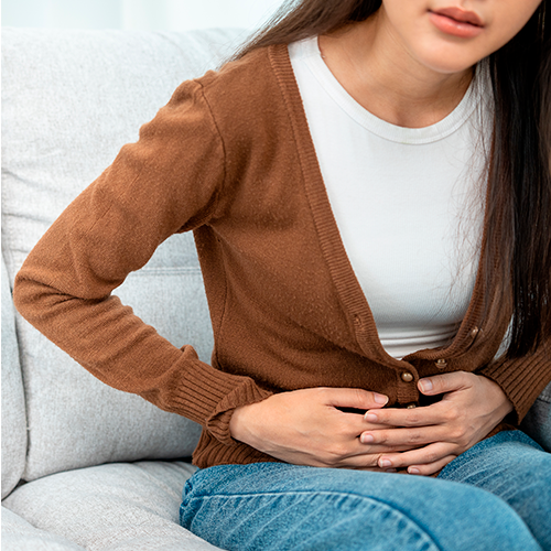 Mulher sentada no sofá segurando o abdômen com as duas mãos, demonstrando desconforto ou dor abdominal.
