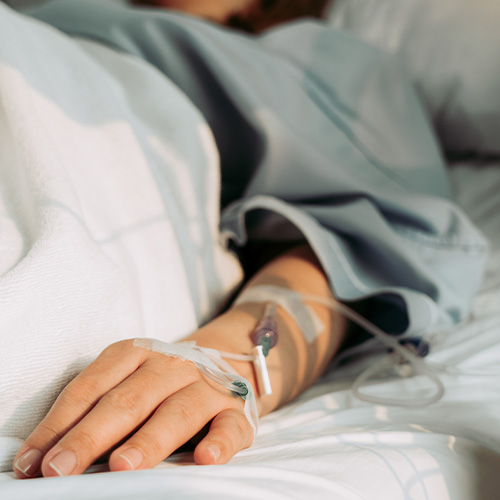 Pessoa deitada em leito hospitalar com acesso venoso no braço, recebendo medicação intravenosa.