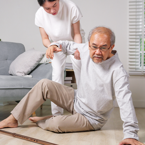 Idoso no chão sendo auxiliado por cuidadora após queda, em ambiente doméstico.