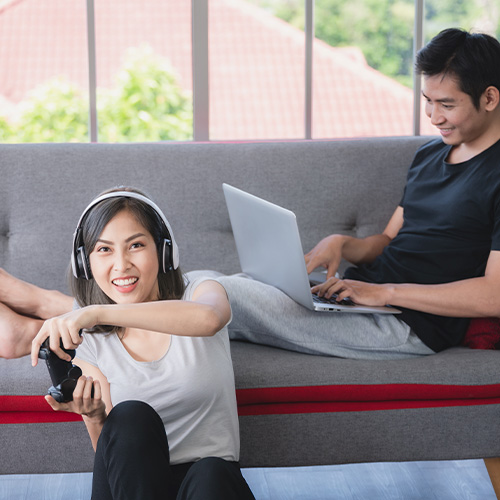 Mulher joga videogame com fones enquanto homem usa notebook no sofá, em ambiente doméstico descontraído.