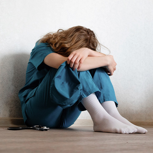 Pessoa sentada no chão abraçando as pernas, com postura retraída em ambiente interno e expressão de tristeza.