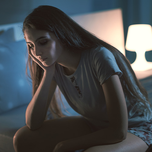 Jovem sentada à noite com expressão cansada, apoiando o rosto na mão em ambiente com luz suave.